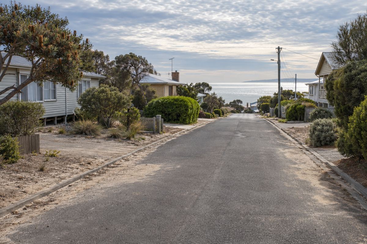 A quiet Rosebud street of weatherboard and fibro holiday houses on stumps beside cream brick post-war homes, tea-tree in the front yards, a sandy nature strip and Port Phillip Bay showing at the end of the road